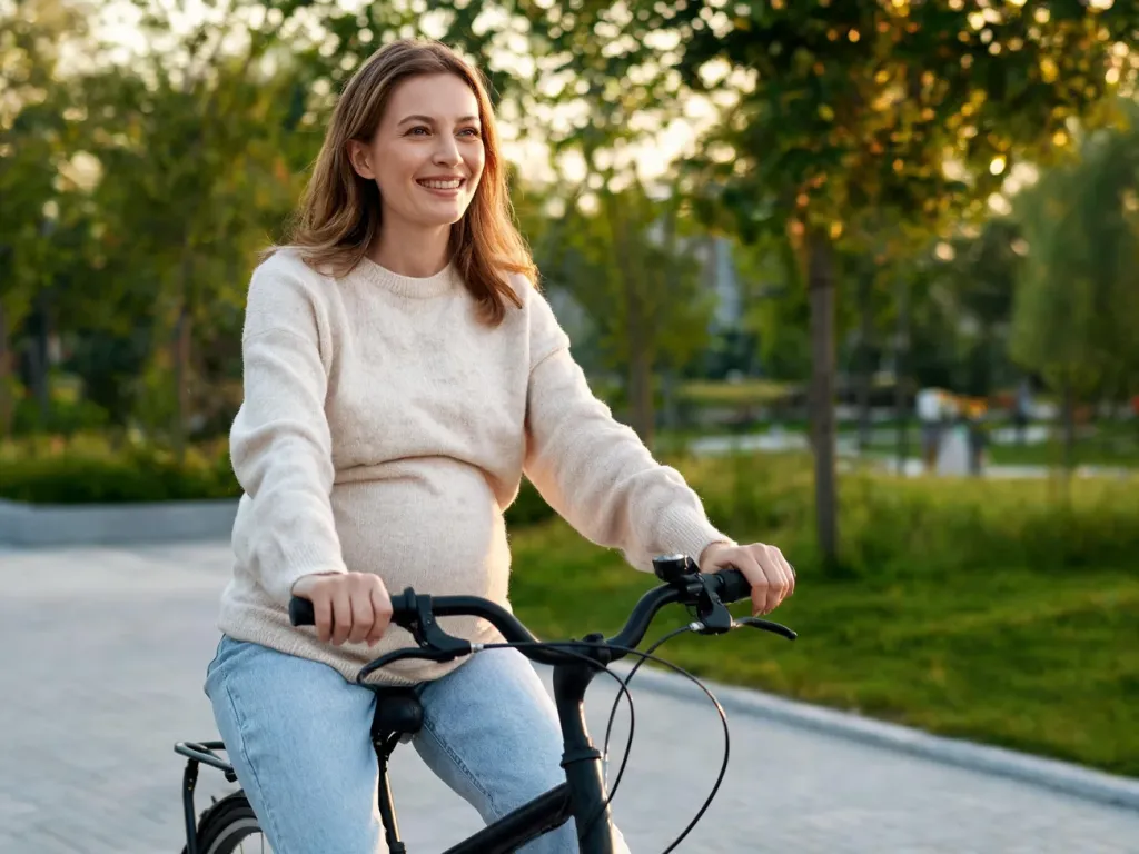 Uśmiechnięta ciężarna kobieta jedzie spokojnie na rowerze po parkowej ścieżce, otoczona zielenią w ciepłym, wieczornym świetle.